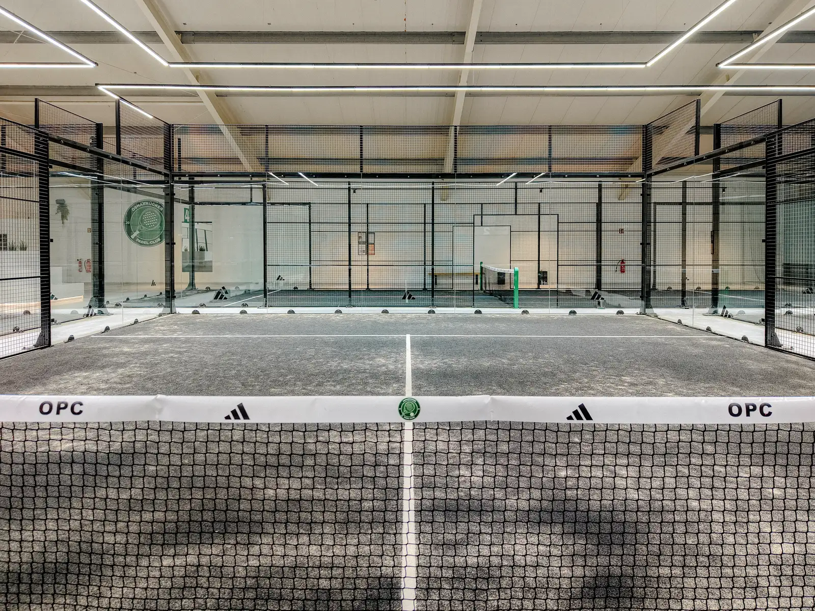 Padelcourt frontal durchs Netz, das Club-Logo an der Wand